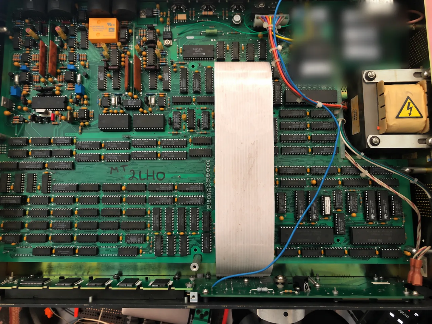 Inside an H3000-D/SE with the top cover removed. Rows of chips cover the board, a wide ribbon cable runs through the center, the power transformer sits on the right and rear panel connectors line the upper edge. Part of the upper right is blurred.